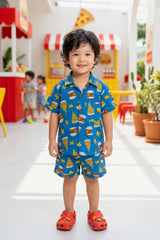 A young boy aged approximately 3–4 years, smiling and wearing the Bebe Unicorn Boys Blue Fast Food Doodle Print Coord Set — a royal blue 100% cotton coord set with bold illustrated burger, pizza and lightning bolt graphic print — photographed in a bright food court themed indoor setting.