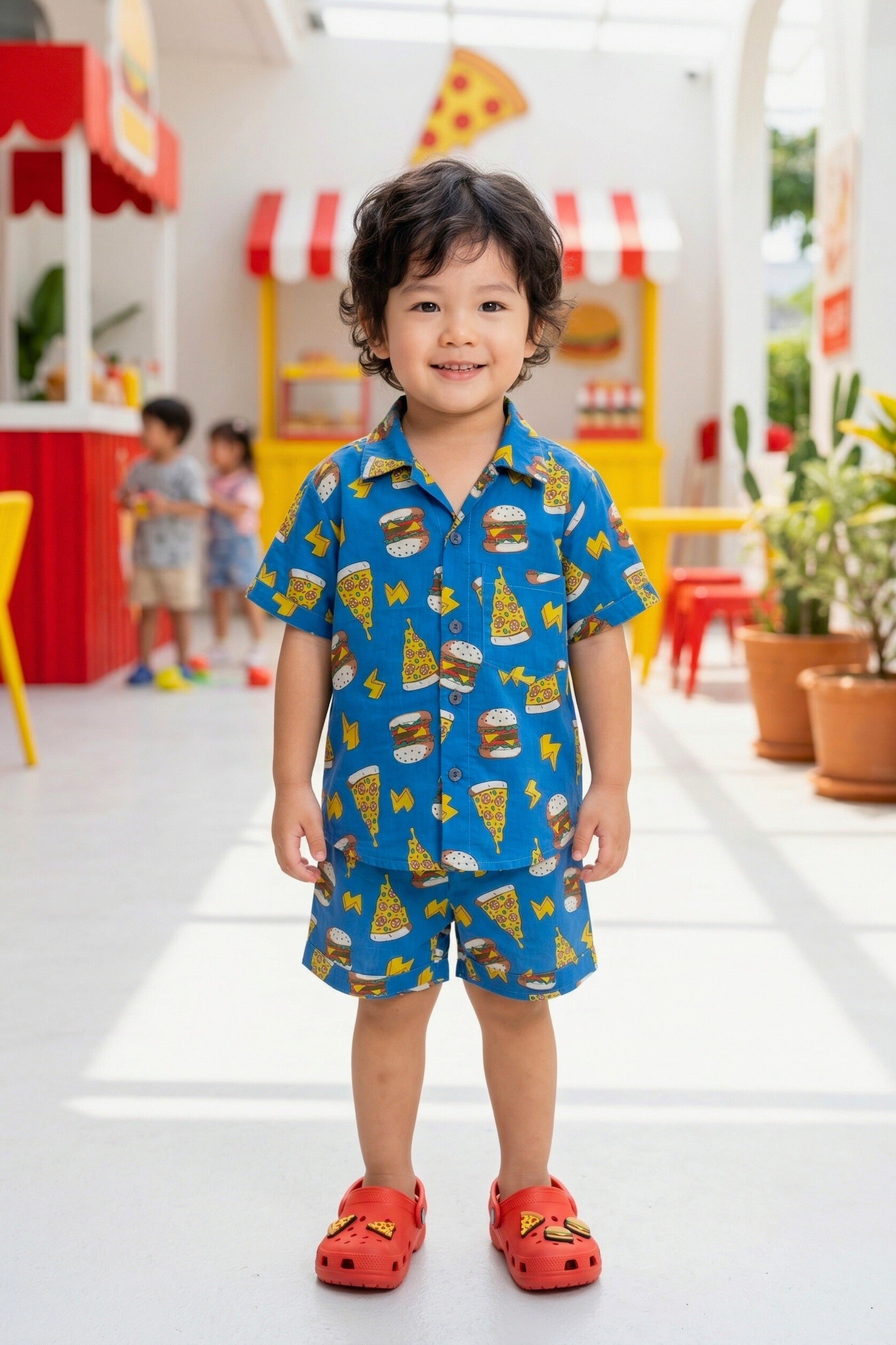 A young boy aged approximately 3–4 years, smiling and wearing the Bebe Unicorn Boys Blue Fast Food Doodle Print Coord Set — a royal blue 100% cotton coord set with bold illustrated burger, pizza and lightning bolt graphic print — photographed in a bright food court themed indoor setting.