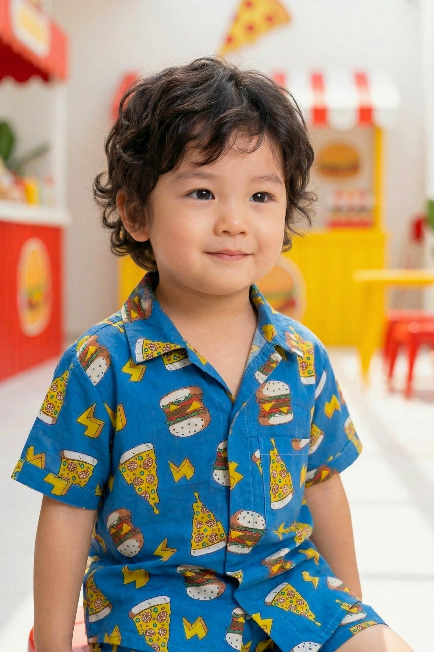 Close-up portrait of a young boy wearing the Bebe Unicorn Boys Blue Fast Food Doodle Print Coord Set, showing the camp collar, coconut shell buttons, chest pocket and bold fast food graphic print on royal blue cotton fabric.