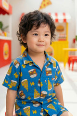 Close-up portrait of a young boy wearing the Bebe Unicorn Boys Blue Fast Food Doodle Print Coord Set, showing the camp collar, coconut shell buttons, chest pocket and bold fast food graphic print on royal blue cotton fabric.
