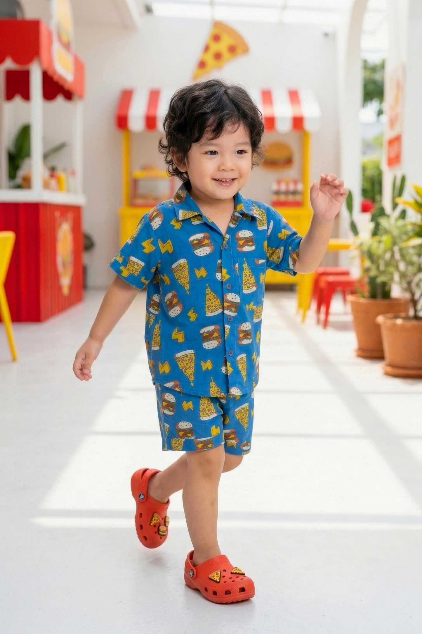 A young boy walking and smiling in the Bebe Unicorn Boys Blue Fast Food Doodle Print 100% Cotton Coord Set — showing the matching shirt and shorts in the graphic burger, pizza and lightning bolt print on royal blue — photographed in a colourful diner-themed indoor setting.