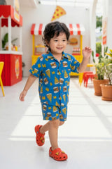 A young boy walking and smiling in the Bebe Unicorn Boys Blue Fast Food Doodle Print 100% Cotton Coord Set — showing the matching shirt and shorts in the graphic burger, pizza and lightning bolt print on royal blue — photographed in a colourful diner-themed indoor setting.