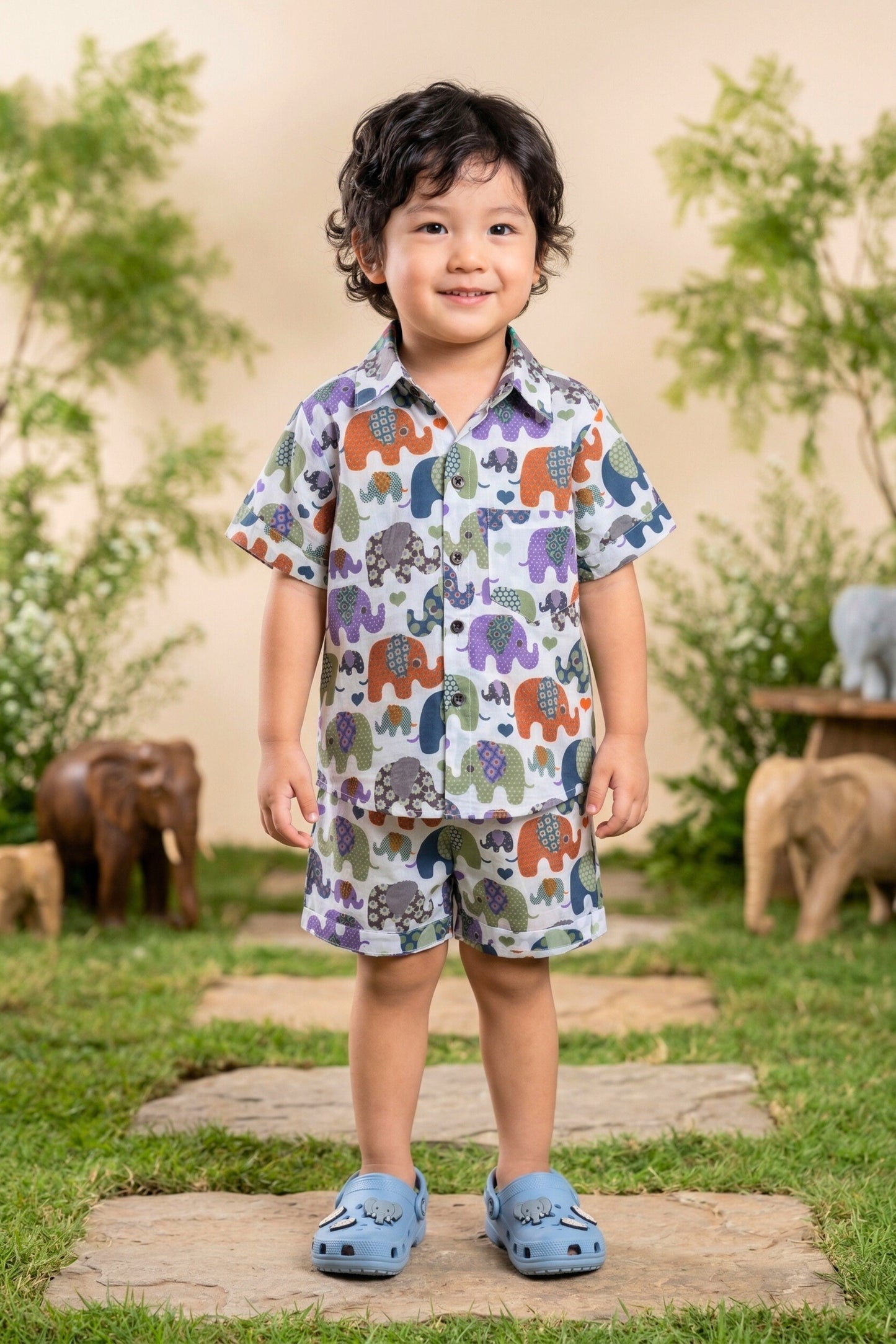 Close-up portrait of a young boy wearing the Bebe Unicorn Boys Multicolour Elephant Parade Print Coord Set, showing the classic shirt collar, coconut shell buttons, chest pocket and the detailed patchwork elephant parade print — six colourful illustrated elephants with unique floral and geometric patterned bodies on white 100% cotton fabric.