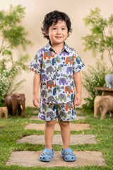 Close-up portrait of a young boy wearing the Bebe Unicorn Boys Multicolour Elephant Parade Print Coord Set, showing the classic shirt collar, coconut shell buttons, chest pocket and the detailed patchwork elephant parade print — six colourful illustrated elephants with unique floral and geometric patterned bodies on white 100% cotton fabric.