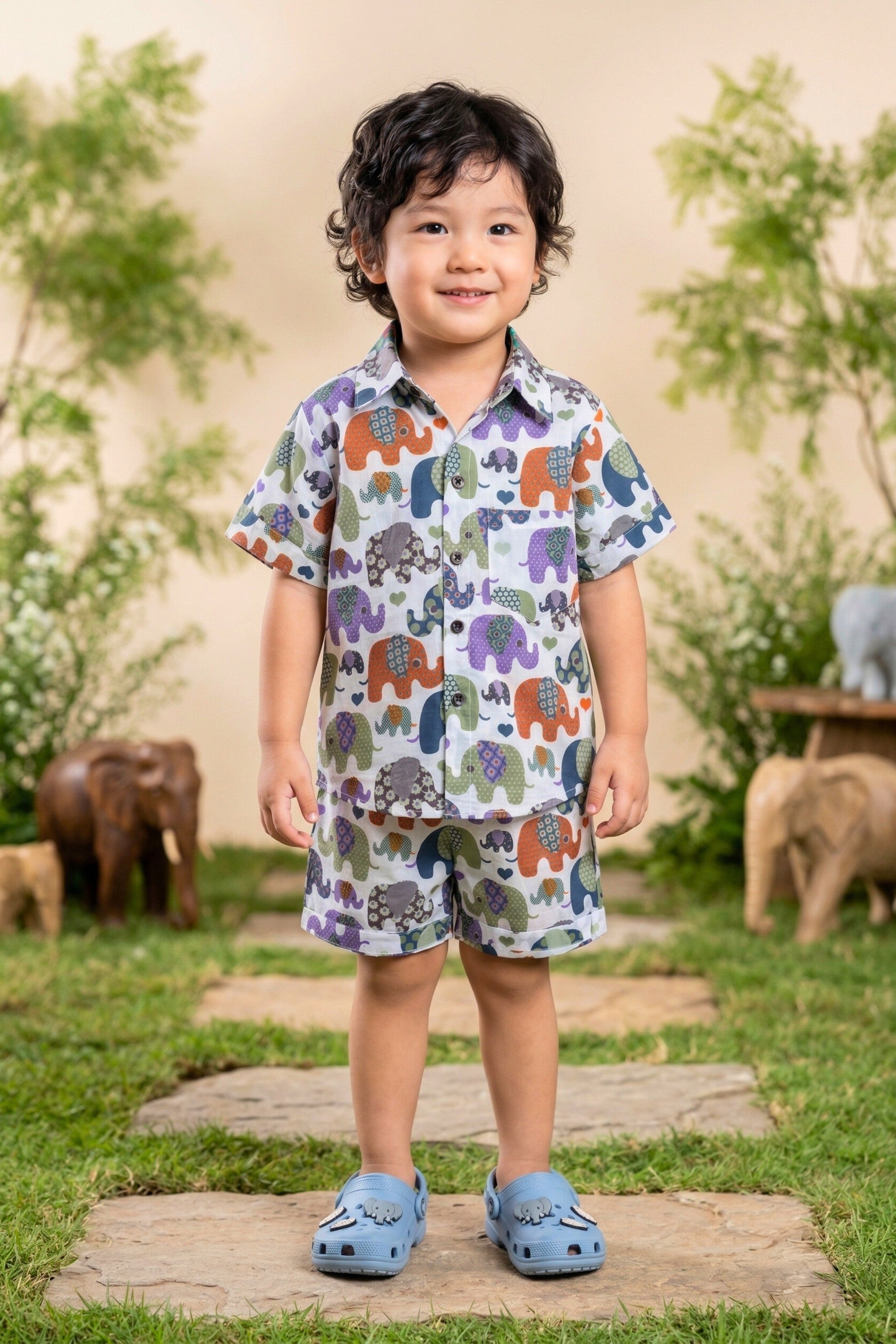 Close-up portrait of a young boy wearing the Bebe Unicorn Boys Multicolour Elephant Parade Print Coord Set, showing the classic shirt collar, coconut shell buttons, chest pocket and the detailed patchwork elephant parade print — six colourful illustrated elephants with unique floral and geometric patterned bodies on white 100% cotton fabric.