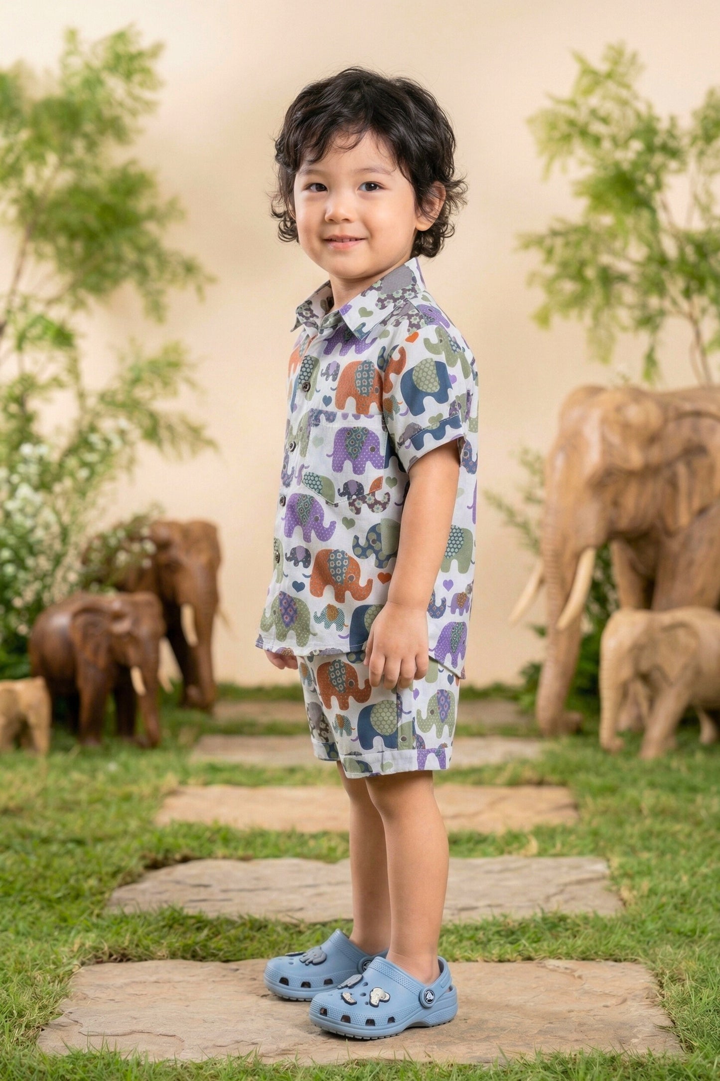 A young boy walking and laughing in the Bebe Unicorn Boys Multicolour Elephant Parade Print 100% Cotton Coord Set — showing the full matching shirt and shorts with the joyful multicolour patchwork elephant parade and scattered hearts — photographed in an outdoor elephant-themed garden setting.