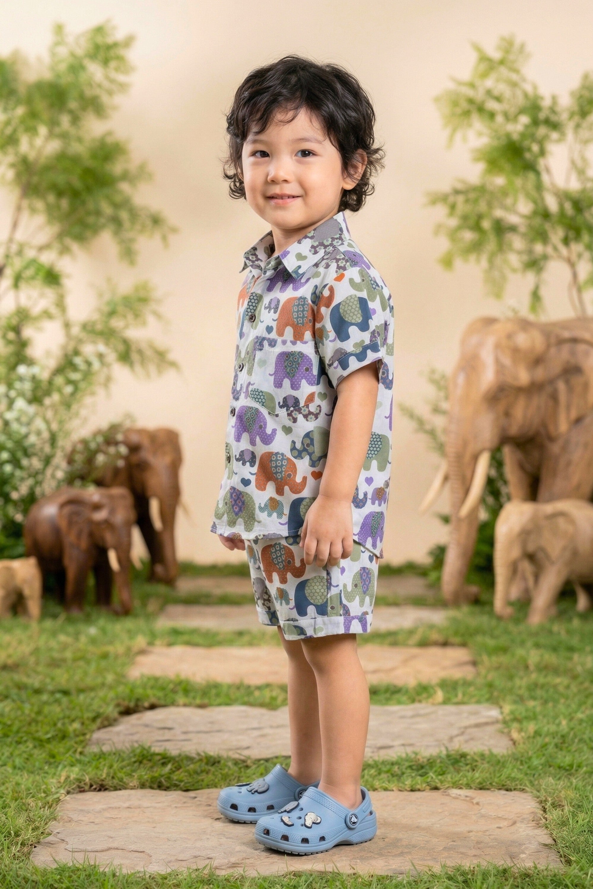 A young boy walking and laughing in the Bebe Unicorn Boys Multicolour Elephant Parade Print 100% Cotton Coord Set — showing the full matching shirt and shorts with the joyful multicolour patchwork elephant parade and scattered hearts — photographed in an outdoor elephant-themed garden setting.