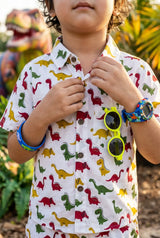 Close-up portrait of a young boy in the Bebe Unicorn Boys Dinosaur Adventure Coord Set — showing the classic collar, short sleeves, functional chest pocket, and detailed colorful dinosaur print (red T-Rex, yellow and green dinos) on white 100% cotton — warm natural light highlighting the pocket detail.