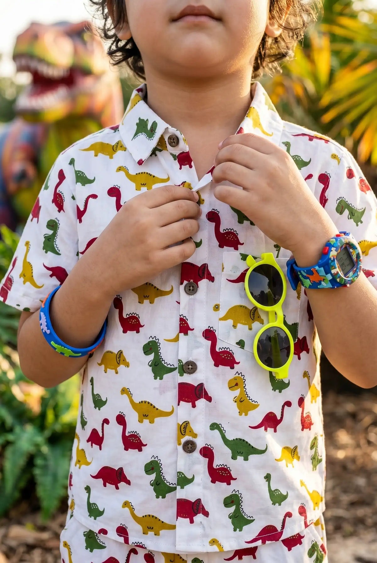 Close-up portrait of a young boy in the Bebe Unicorn Boys Dinosaur Adventure Coord Set — showing the classic collar, short sleeves, functional chest pocket, and detailed colorful dinosaur print (red T-Rex, yellow and green dinos) on white 100% cotton — warm natural light highlighting the pocket detail.