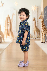 Close-up portrait of a young boy wearing the Bebe Unicorn Boys Navy Space Giraffe Print Coord Set, showing the classic shirt collar, coconut shell buttons, chest pocket and the detailed space giraffe print — golden giraffes floating alongside teal, pink and amber planets on a deep navy cotton fabric.
