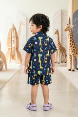 A young boy walking and smiling in the Bebe Unicorn Boys Navy Space Giraffe Print 100% Cotton Coord Set — showing the full matching shirt and shorts with the whimsical space giraffe, planet and rocket print — photographed in a pastel space-themed studio with giraffe and planet props in the background.