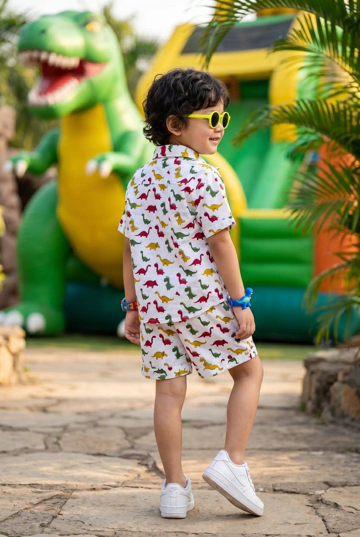 Close-up fabric detail of the colorful dinosaur print on the Bebe Unicorn boys adventure coord set — vibrant red T-Rex, yellow dinosaurs, green dinosaurs, and multi-color dino mix on white 100% cotton base — showcasing the playful, high-energy primary colors and cartoon aesthetic for children aged 2–8 years.