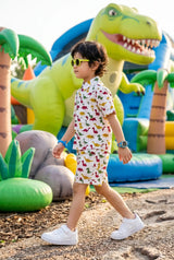 Full length view of a young boy walking in the Bebe Unicorn Boys Colorful Dinosaur Adventure Coord Set — showing the complete outfit silhouette with matching shirt and shorts, bright colorful dinosaur print throughout, and the side pockets visible on the shorts — outdoor adventure setting.