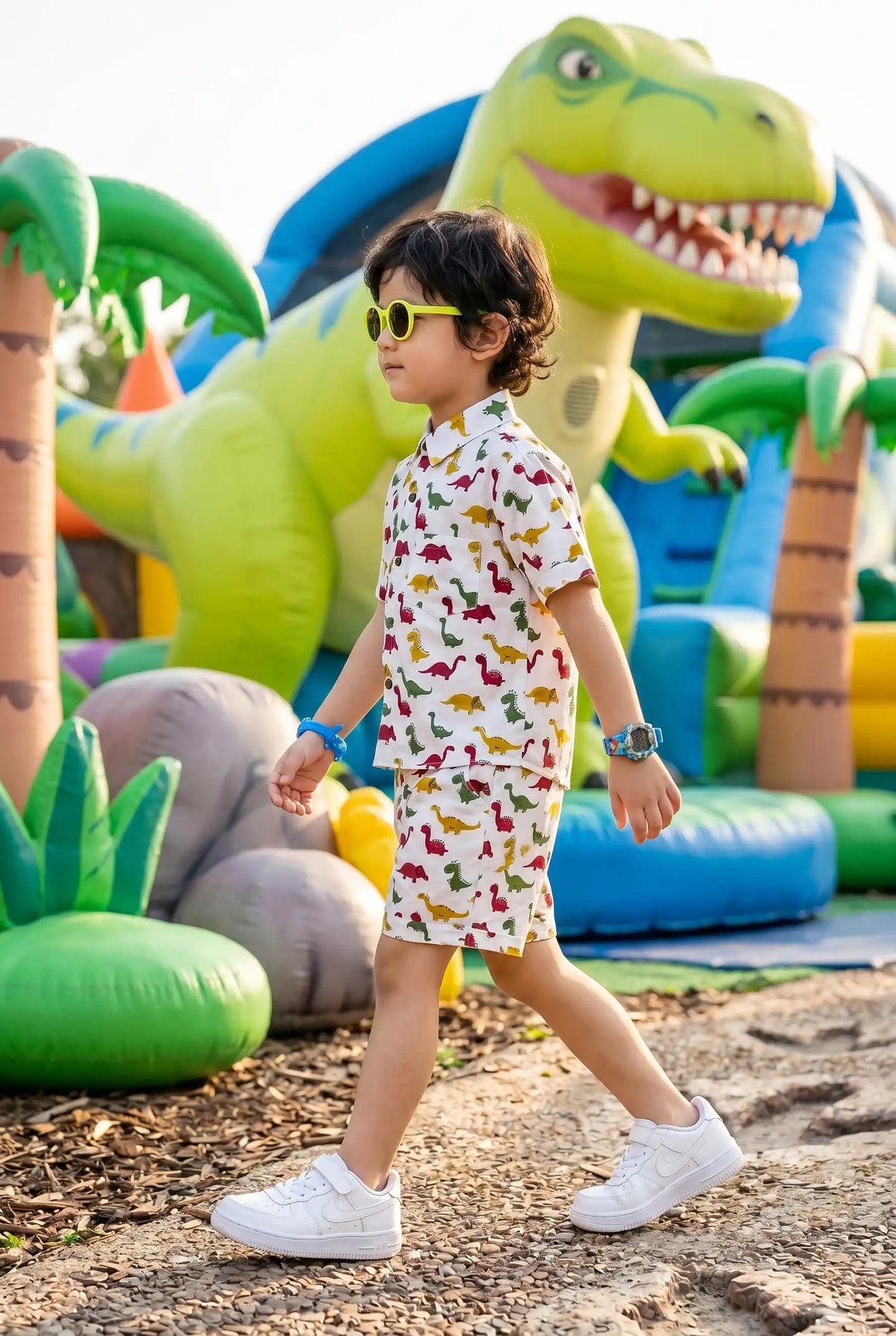 Full length view of a young boy walking in the Bebe Unicorn Boys Colorful Dinosaur Adventure Coord Set — showing the complete outfit silhouette with matching shirt and shorts, bright colorful dinosaur print throughout, and the side pockets visible on the shorts — outdoor adventure setting.