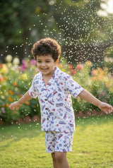 Image 2 — Full length front view:
Full length front view of a young boy wearing the Bebe Unicorn Boys Surf & Beach Woody Car Print Linen Coord Set — showing the camp collar shirt with coconut shell buttons and chest pocket paired with matching linen shorts — photographed in a garden with natural green grass and golden hour light.