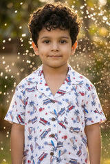 Image 1 — Hero close-up / portrait:
A young Indian boy aged approximately 5–6 years, smiling and wearing the Bebe Unicorn Boys Surf & Beach Woody Car Print 100% Linen Coord Set — a white linen shirt with red and blue vintage surf car, palm tree and hibiscus illustrations — photographed outdoors in golden hour light with a water sprinkler bokeh background.