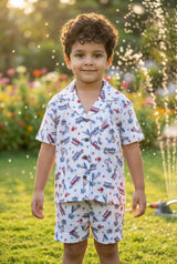 Image 3 — Side / three-quarter view:
Side view of a young boy in the Bebe Unicorn Boys Surf & Beach Woody Car Print 100% Linen Coord Set, showing the linen fabric texture, camp collar and matching printed shorts — outdoor garden setting with warm natural lighting.